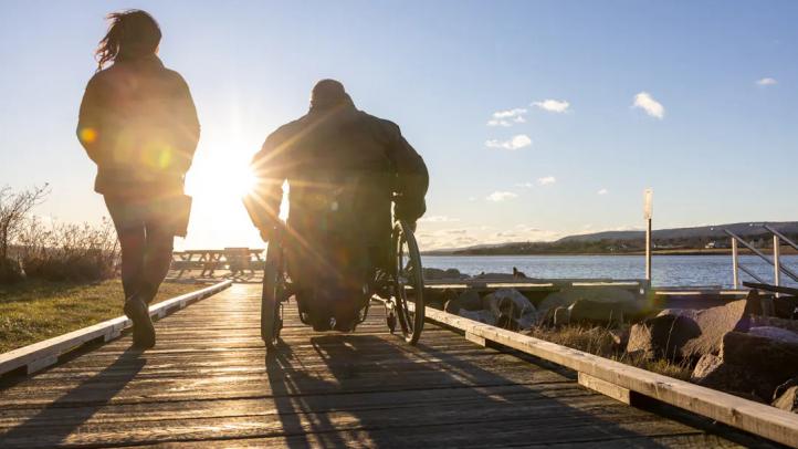 Two people, one walking and one in a wheelchair, on a boardwalk heading into the sunset near the beach